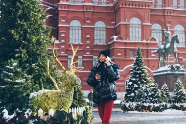 Siyah ceketli, gülümseyen bir kadın. Parlayan caddede yürürken cep telefonuyla sohbet ediyor. Yeni yıl için köknar süslemeleri yapıyor.