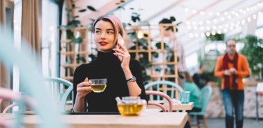 Şık, mutlu, pembe saçlı, günlük kıyafetleri olan, akıllı telefondan konuşan ve hipster kafeteryasında oturmuş çay içen bir kadın.
