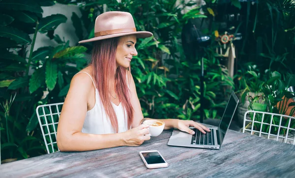 在温暖的夏日 绿色花园的工作场所 一位戴着帽子 头戴白色上衣的漂亮女性博主在观看手提电脑屏幕时的侧影 — 图库照片