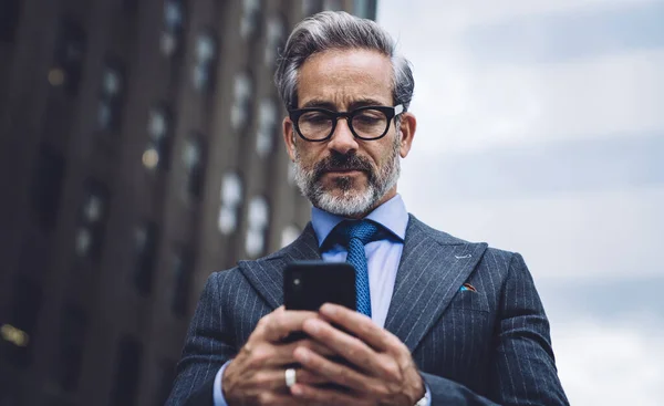 From below concentrated bearded middle aged man in formal suit and glasses typing on mobile phone in downtown area of New York