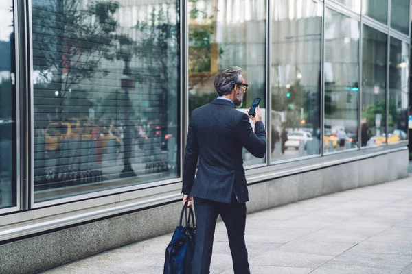 Back view of successful middle aged man in stylish dark business suit walking down city street with smartphone and bag in New York