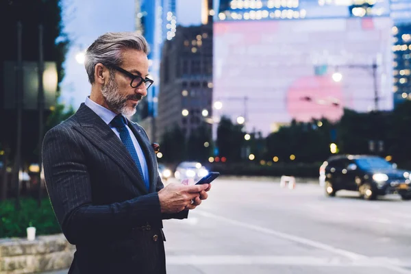 Side view of middle aged male in glasses and business suit typing on smartphone and standing against evening New York road