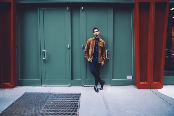 Handsome young man with hand in pocket, legs crossed and glasses on shirt in trendy clothes leaning on green entrance while looking at camera on New York street