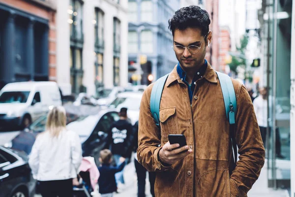 一位英俊的黑发男性游客穿着棕色绒面革夹克 背着蓝色背包和眼镜 一边听着耳机里的音乐 一边在纽约街头用手机浏览 — 图库照片