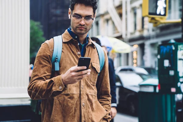 身穿褐色羊皮夹克 背着背包和耳机 戴着眼镜的英俊的西班牙裔男性 站在纽约街上的红绿灯旁 用手机发短信 — 图库照片