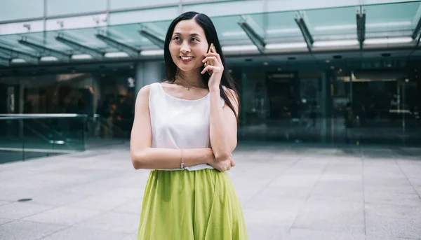 Beautiful Asian woman in white top and lime skirt talking on phone while standing in front of modern office building