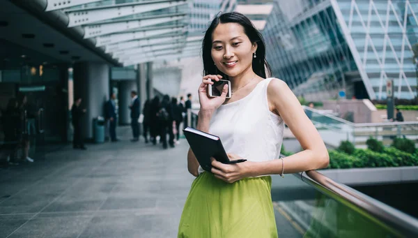 現代的なスマートフォンやノートブックの笑顔と市内のオフィスビルの外のテラスの手すりにもたれて美しいアジアの女性起業家 — ストック写真
