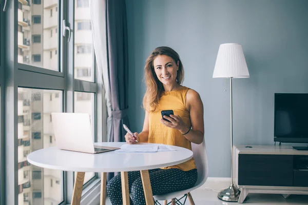 Glad young woman in yellow sleeveless blouse sitting at white round table with laptop and paper typing on smartphone at home and looking at camera