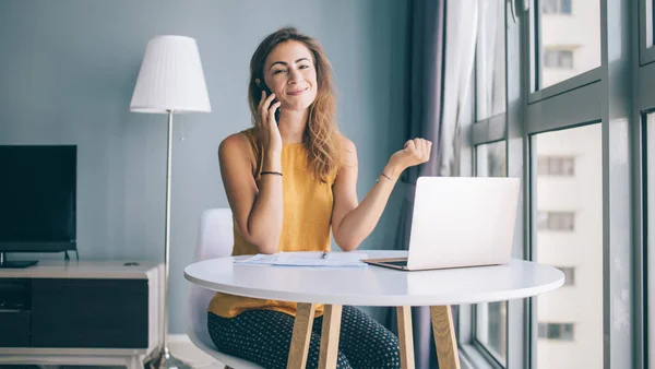 Smiling young woman in yellow sleeveless blouse sitting at white round table with paper and laptop communicating on smartphone at home looking at camera