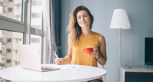 Happy young attractive woman in yellow sleeveless blouse sitting at white round table with laptop and red mug writing on paper at home and looking at camera