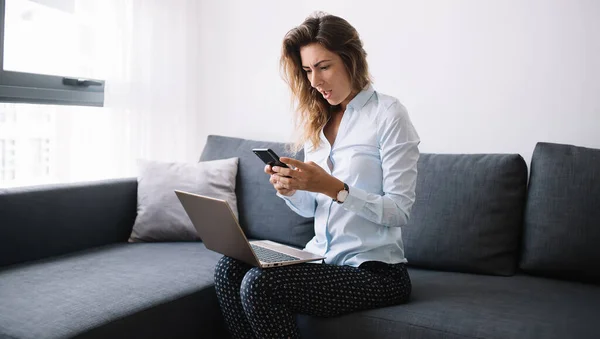 Jovem Atordoada Camisa Usando Telefone Celular Sentado Sofá Com Laptop — Fotografia de Stock