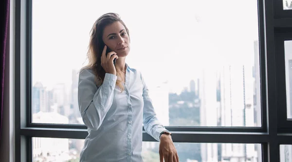 穿着白衬衫的成功成年女性在电话交谈时 靠在大窗户上 从城市的视野看问题 — 图库照片