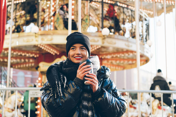 Toothy smiling young female with red lips wearing black winter hat standing by festive New Year carousel with coffee to go while dreaming and looking away