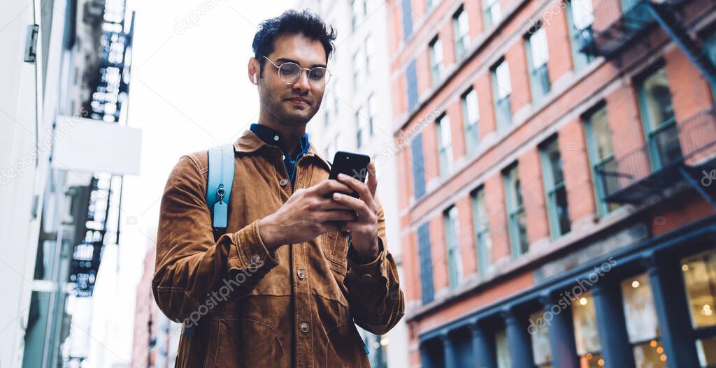 From below handsome stylish Hispanic male in eyeglasses wearing brown suede jacket with earphones chatting on cellphone while standing on sidewalk in New York city between office buildings