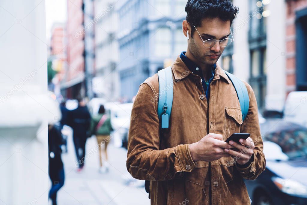 Serious handsome male in eyeglasses wearing brown suede jacket with backpack and earphones messaging on cellphone while going down street in business district of New York city