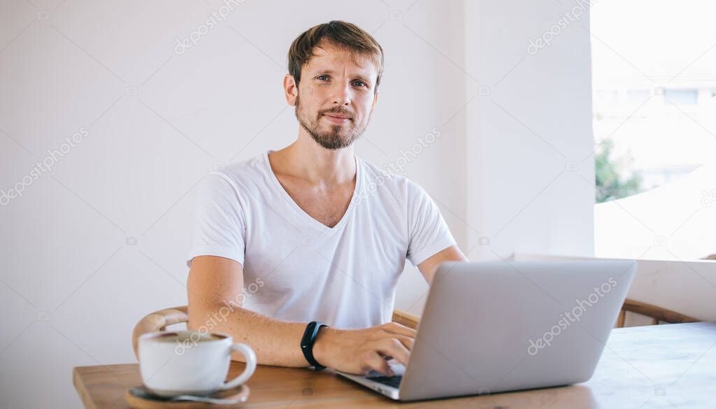 Portrait of handsome male freelancer in looking at camera while working remotely at modern laptop computer connected to wireless free 4g internet sitting in coffee shop, concept of distance job
