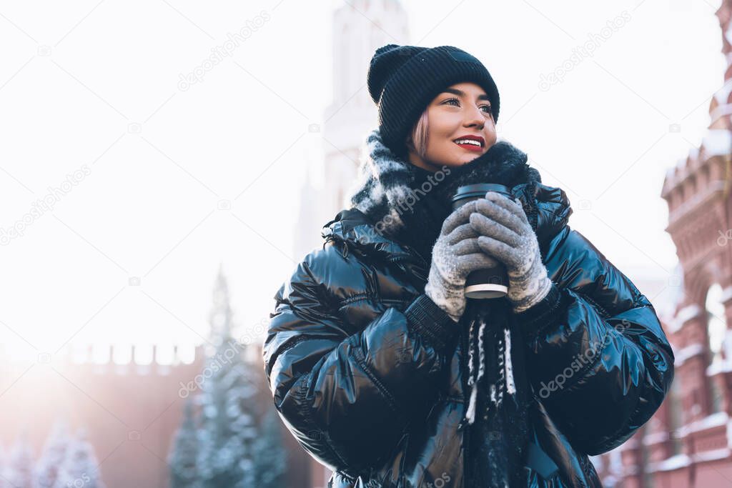 Happy young woman in comfortable oversize winter jacket resting near historical buildings with warm coffee in plastic cup on blurred background and looking away