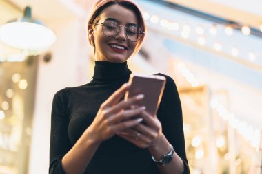 Siyah ve kırmızı gündelik giysiler içindeki çekici genç kadının yumuşak odağı ve arka planda bulanık lambaların üzerinde kısa saçlı akıllı telefonun gezinmesi. 