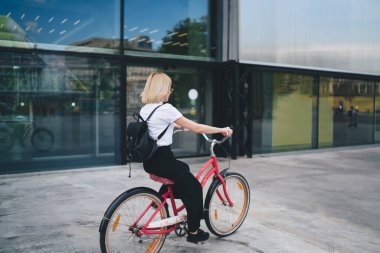 Sokakta bisiklet sürerken kırmızı çerçeveli siyah sırt çantalı sarışın kız öğrencinin arka planı.