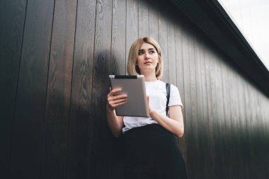 Ahşap duvara yaslanıp tablet kullanırken dalgın ve çekici bir kız öğrencinin dalgın bakışları.