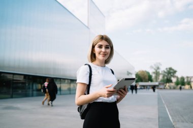 Günlük kıyafetlerle gezen neşeli kadın, şehir caddesinde ofis binasının yanında durup kameraya gülümserken zevkle tableti karıştırıyor.