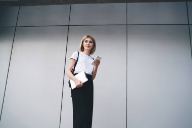 Sıradan kıyafetli, siyah sırt çantalı, tablet taşıyan ve sokak binasına karşı akıllı telefon kullanan dalgın, sarışın bir kadın. 