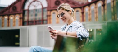 Binanın bulanık arka planında yeşil çalıların arkasında otururken gülümseyen ve akıllı telefonlara bakan sarışının yan görüntüsü