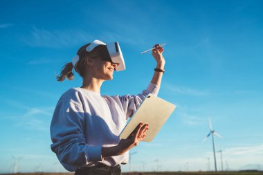 VR kulaklıklı sıradan gülümseyen kadın dokunmatik programla sanal gerçeklikte oynuyor ve sahada Stylus 'la el kol hareketleri yapıyor mavi gökyüzü arka planında yel değirmenleri var.