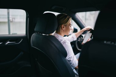 Beyaz tişörtlü, açık renk saçlı bir kadın, siyah bir araçta oturuyor. Direksiyonda oturuyor ve yüzünde gülümseme olan akıllı telefon ekranına bakıyor.