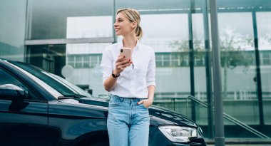 Siyah arabaya yaslanıp akıllı telefondan mesajlaşırken günlük kıyafetlerle gülümseyen neşeli sarışın kadın sürücü.