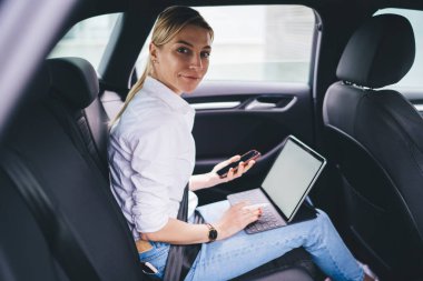 Gülümseyen kadın yönetici emniyet kemeriyle arabanın arka koltuğuna bağlanmış, akıllı telefon tutuyor ve kameraya bakarken tablet klavyeye yazı yazıyor.