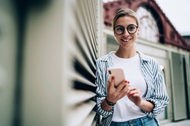 Yuvarlak gözlüklü ve çizgili tişörtlü neşeli sarışın kadın cep telefonu kullanıyor ve pencereye yaslanırken kapalı bir külüstürle kameraya bakıyor.