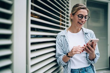 Neşeli, genç, güzel bir bayan günlük kıyafetler ve moda gözlükler takıp gülüyor, akıllı telefonlara bakıyor ve bina yakınlarında komik videolar seyrediyor.