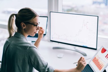 Modern ofisin penceresinin yanında bilgisayar ve tabletle çalışan çağdaş, zeki iş kadınının yan görünüşü 