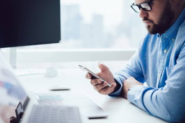 Gözlüklü yetişkin bir adamın bilgisayar masasında dizüstü bilgisayarla oturması ve gün ışığında cep telefonuyla sörf yapması.