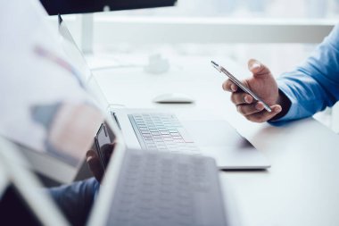 Modern ışık ofisindeki beyaz masada tablet ve dizüstü bilgisayarla otururken mavi gömlekli yüzsüz iş adamı akıllı telefon sörfü yapıyor. 