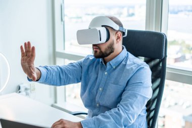 Yetişkin sakallı bir adam açık beyaz bir ofiste masada oturuyor ve VR gözlükleri projesinde çalışırken elini uzatıyor.