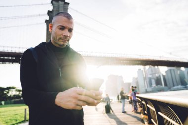 Siyah spor giyim giymiş, keyifli atletik genç adam şehir köprüsünde arka plandaki metal çitlerde akıllı telefonuna dokunuyor.