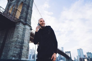 Gülümseyen, kısa saçlı, siyah giysili genç adam şehir planında güpegündüz köprüde akıllı telefonla iletişim kuruyor.