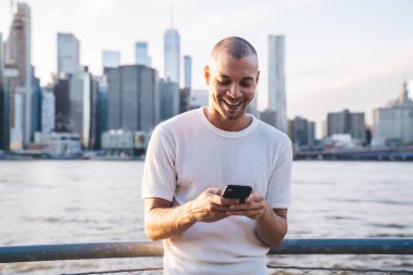 Beyaz tişörtlü genç, kısa saçlı, gülen adam gökdelenlerin arka planında çitlere yaslanmış ve akıllı telefonuyla yazı yazıyor.