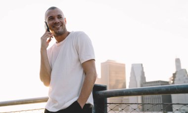 Aşağıdaki beyaz tişörtlü, kısa saçlı adamdan tırabzana yaslanmış ve telefonda gülümseyen.