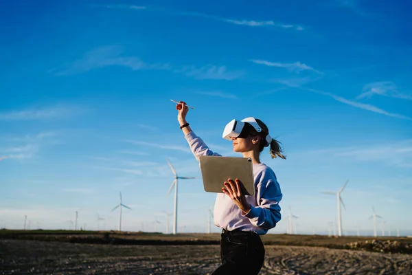 Side view of lady in VR headset studying digital virtual reality space with touchpad program and painting with pencil in air being in big field with windmills on bright blue sky background