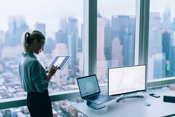 带着自信的时尚女商人站在当代办公室 靠窗的台式机面对城市景观时使用平板电脑的侧视图 — 图库照片