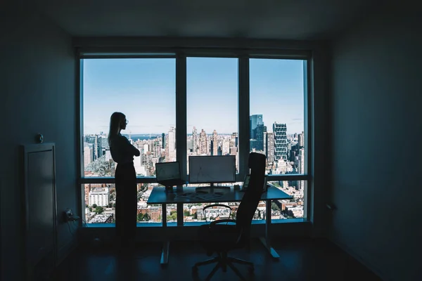 Side view of female silhouette standing near computer desktop and looking pensively away in window against contemporary city view behind