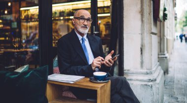 Gözlüklü, takım elbiseli, akıllı telefonlu yaşlı bir adam cama yaslanmış ve kahve fincanıyla yan yana oturuyor.