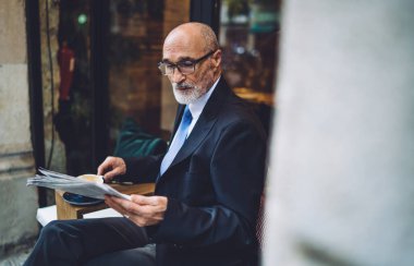 Sakallı, kahve bardaklı, takım elbiseli, beyaz duvarın yanında oturan ve gazete okuyan yaşlı adamın yan görüntüsü.