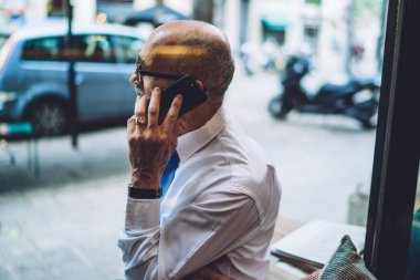 Beyaz tişörtlü, kısa saçlı yaşlı işadamının yan görüntüsü şehir sokaklarının ve yolların arka planında oturup telefonla konuşması.