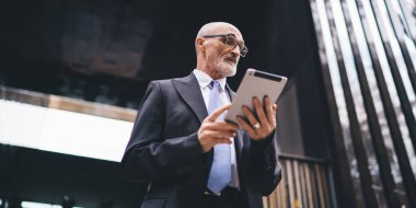 Resmi kıyafetli, gözlüklü ve beyaz sakallı yaşlı, yakışıklı, kel bir erkekten. İş merkezinin önündeki tabletlere göz atıyorlar.