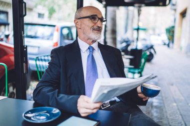 Caddedeki kafede gri sakallı, mavi kravatlı, kahve fincanı ve şehir arka planında gazetesi olan neşeli, son sınıf öğrencisi.