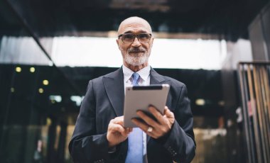 Yakışıklı, sakallı, kel, resmi kıyafetli ve gözlüklü bir adam iş merkezinin önünde kameraya bakıyor.